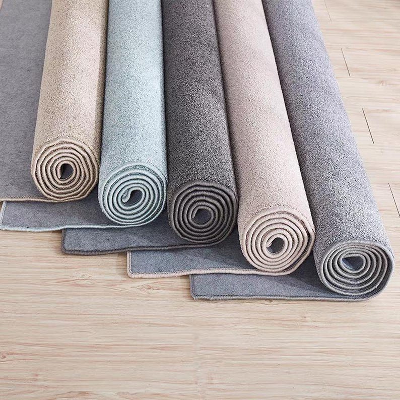 Puede incluir: Alfombras enrolladas en tonos beige, gris y azul, dispuestas sobre un suelo de madera clara. Se aprecia la textura y la variedad de colores de las alfombras. Los bordes de las alfombras son visibles, mostrando su construcci&oacute;n.