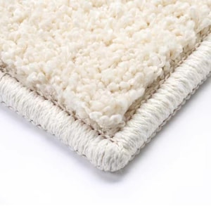 Puede incluir: Primer plano de una alfombra de felpa color crema con un pelo grueso y texturizado. La alfombra tiene un borde tejido en un color crema a juego, creando un borde limpio y acabado. La textura suave y esponjosa sugiere comodidad y calidez.