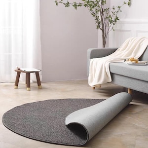 Puede incluir: Una alfombra semicircular gris oscuro con una superficie texturizada. La alfombra est&aacute; parcialmente enrollada, revelando una parte inferior gris claro. La alfombra est&aacute; sobre un suelo marr&oacute;n claro en un sal&oacute;n. Un peque&ntilde;o taburete de madera y un sof&aacute; gris est&aacute;n en el fondo.