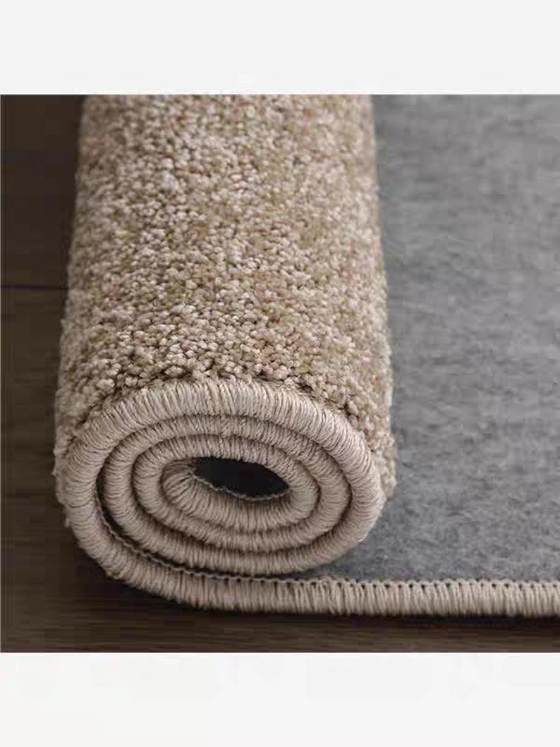 Puede incluir: Alfombra beige enrollada con una superficie texturizada, que revela un reverso gris. El borde de la alfombra est&aacute; cuidadosamente terminado con un borde beige a juego. La imagen resalta la textura y el color afelpado de la alfombra.