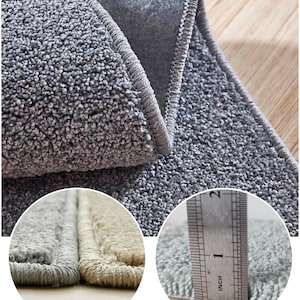 Puede incluir: Primer plano de una alfombra gris y afelpada con borde enrollado. La alfombra tiene una superficie texturizada y un pelo grueso. Una regla muestra que la alfombra tiene aproximadamente 2,5 cm de grosor. La alfombra es de color neutro.