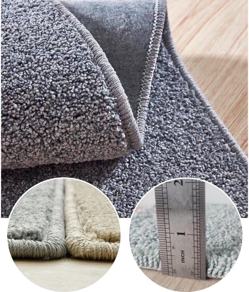 Puede incluir: Primer plano de una alfombra gris y afelpada con borde enrollado. La alfombra tiene una superficie texturizada y un pelo grueso. Una regla muestra que la alfombra tiene aproximadamente 2,5 cm de grosor. La alfombra es de color neutro.