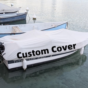 Puede incluir: Una lona blanca con el texto "Custom Cover" en una pequeña embarcación. La embarcación está en el agua cerca de otras embarcaciones. La embarcación tiene un motor negro y defensas blancas.