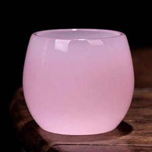 May include: A small, round, pink glass vase with a smooth, glossy finish. The vase has a wide opening and a rounded body. The background is dark, highlighting the soft pink color of the glass. The vase is sitting on a wooden surface.