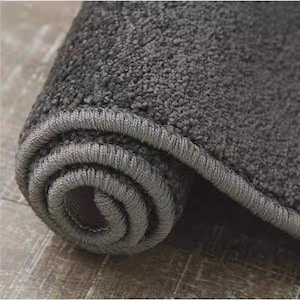 Puede incluir: Alfombra gris carb&oacute;n enrollada, con una superficie texturizada y un borde gris oscuro. La textura afelpada de la alfombra es visible en el primer plano, destacando su suavidad. La forma enrollada sugiere un f&aacute;cil almacenamiento o transporte.