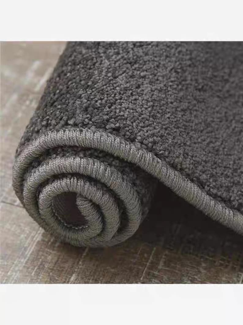 Puede incluir: Alfombra gris carb&oacute;n enrollada, con una superficie texturizada y un borde gris oscuro. La textura afelpada de la alfombra es visible en el primer plano, destacando su suavidad. La forma enrollada sugiere un f&aacute;cil almacenamiento o transporte.