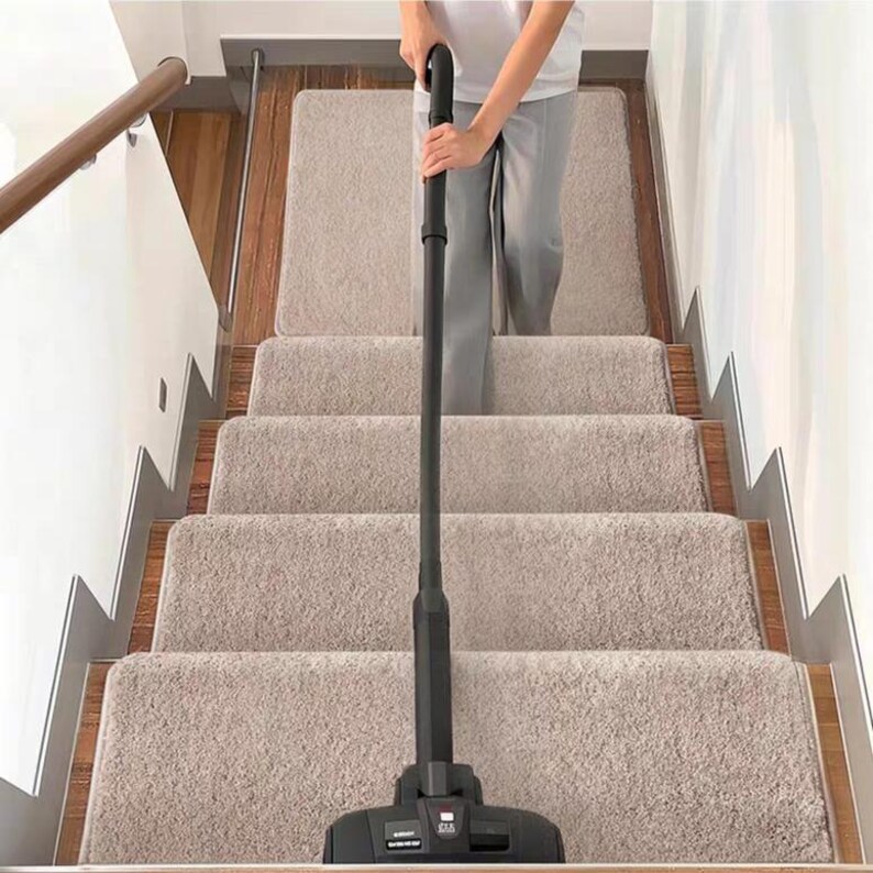 Puede incluir: Una aspiradora negra que se utiliza en una escalera alfombrada. Las escaleras tienen alfombras beige y molduras de madera. La persona que usa la aspiradora lleva pantalones grises y una camisa blanca.