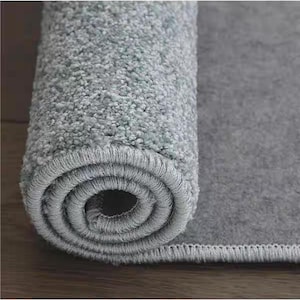 Puede incluir: Alfombra gris claro enrollada, con una superficie texturizada. El borde de la alfombra es visible, mostrando un borde cuidadosamente cosido. La secci&oacute;n enrollada revela el grosor y la construcci&oacute;n de la alfombra, adecuada para la decoraci&oacute;n del hogar.