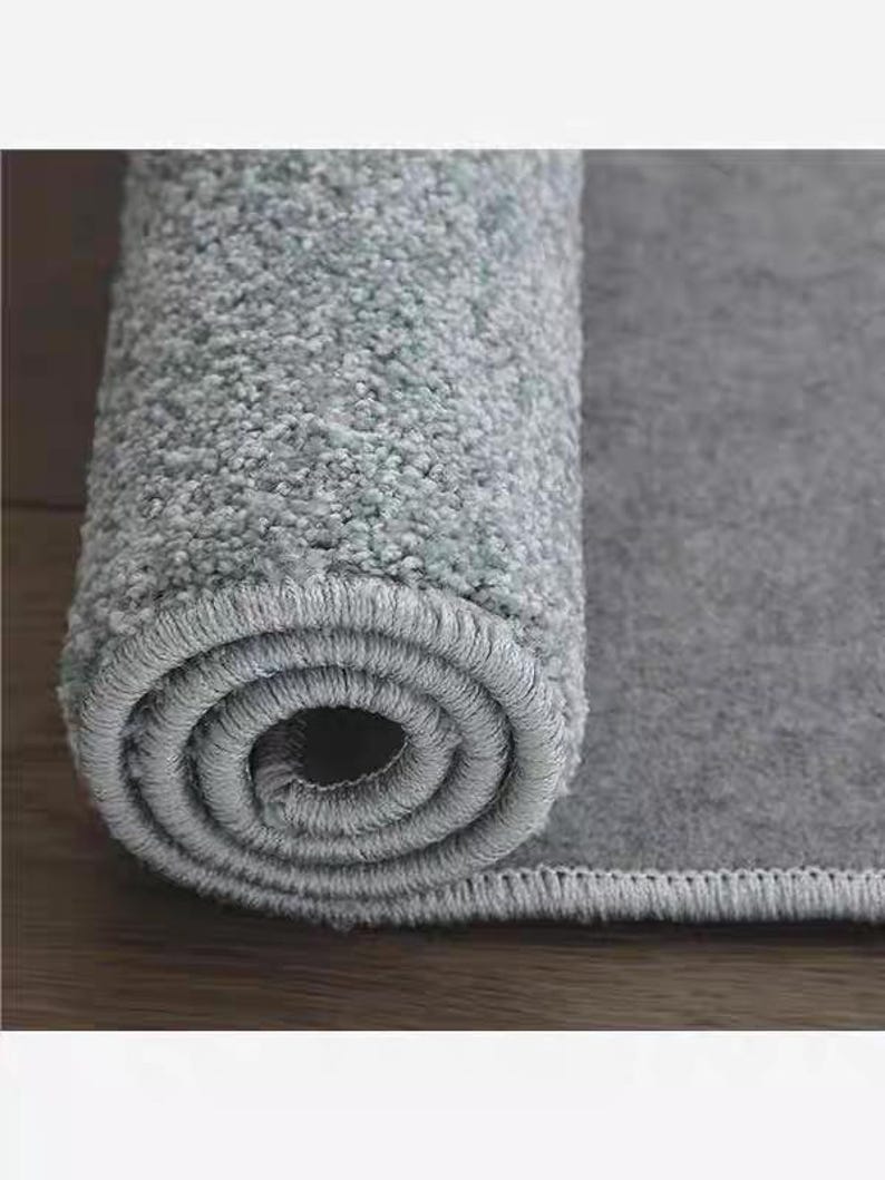 Puede incluir: Alfombra gris claro enrollada, con una superficie texturizada. El borde de la alfombra es visible, mostrando un borde cuidadosamente cosido. La secci&oacute;n enrollada revela el grosor y la construcci&oacute;n de la alfombra, adecuada para la decoraci&oacute;n del hogar.
