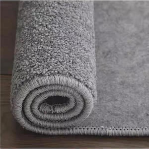 Puede incluir: Una alfombra gris enrollada, con una superficie texturizada y suave. El borde de la alfombra tiene un borde cosido. La secci&oacute;n enrollada revela el grosor y la construcci&oacute;n de la alfombra, adecuada para la decoraci&oacute;n del hogar.