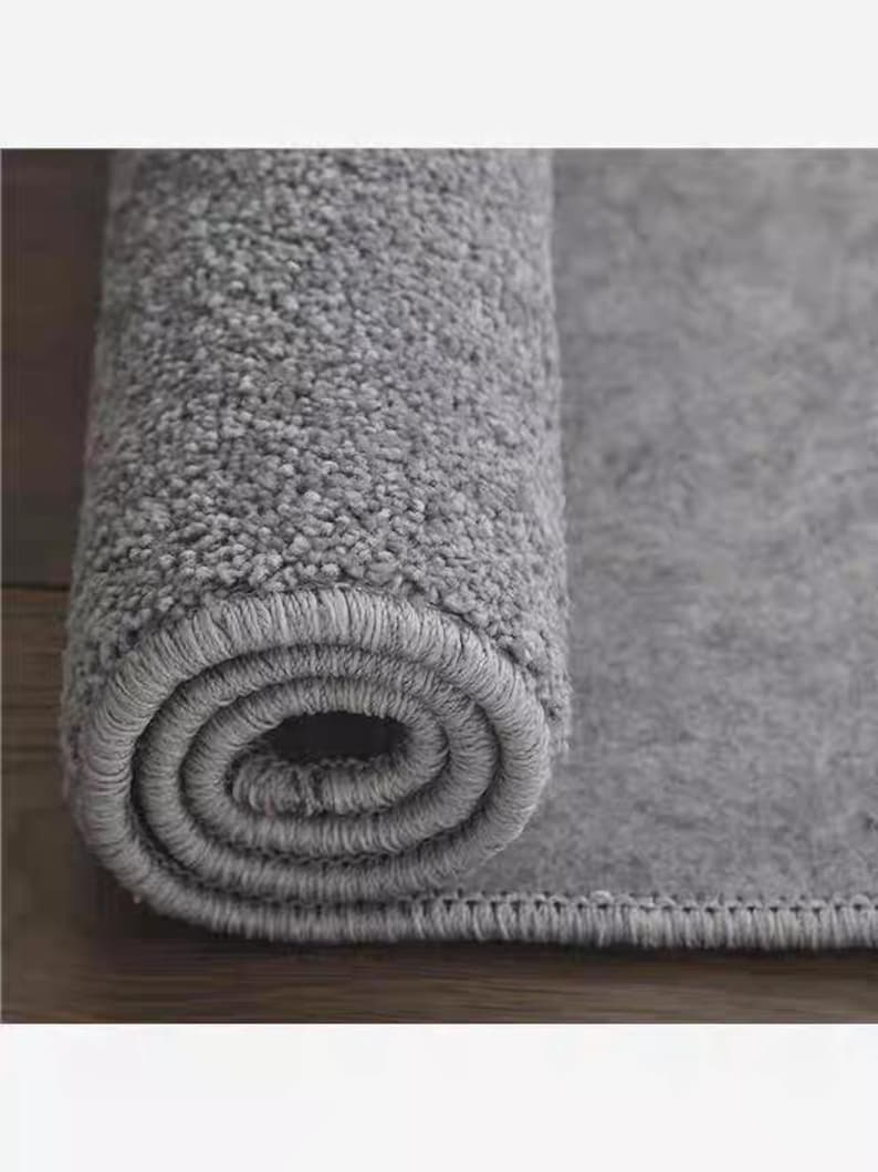 Puede incluir: Una alfombra gris enrollada, con una superficie texturizada y suave. El borde de la alfombra tiene un borde cosido. La secci&oacute;n enrollada revela el grosor y la construcci&oacute;n de la alfombra, adecuada para la decoraci&oacute;n del hogar.
