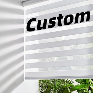 Peut inclure: Stores de fenêtre à rayures horizontales blanches et grises avec le mot "Custom" en grandes lettres noires. Les stores sont partiellement ouverts, révélant une vue de plantes vertes à l'extérieur. Les stores sont montés sur un mur blanc.