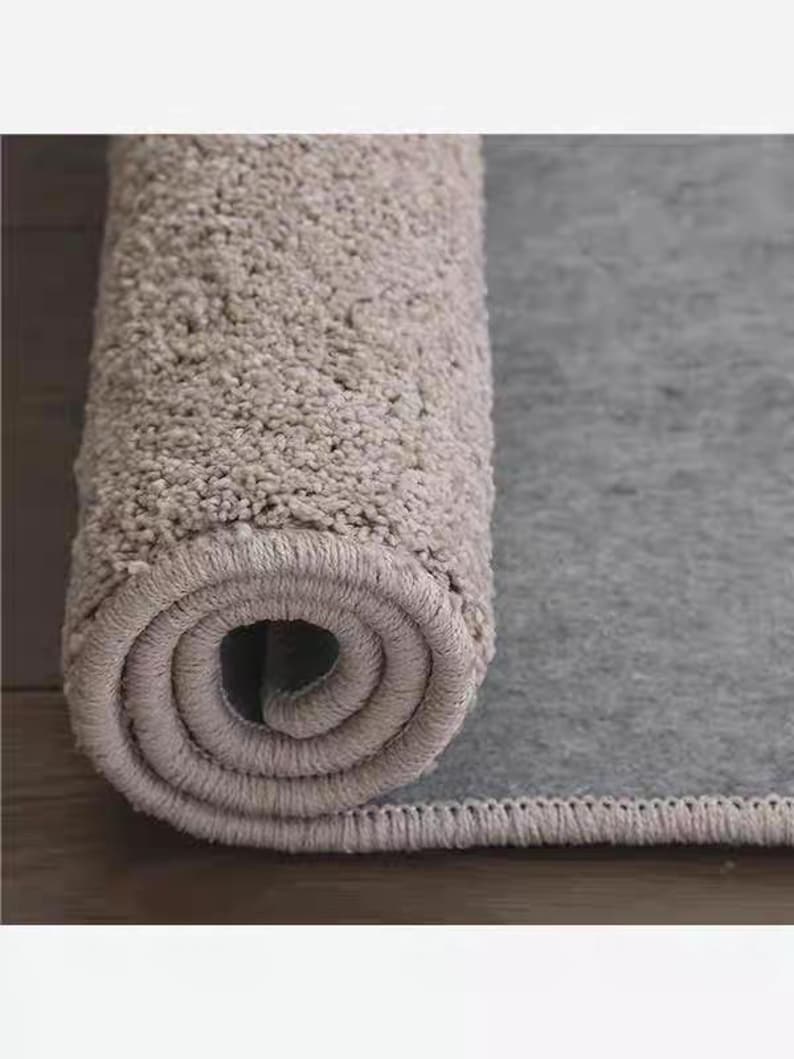 Puede incluir: Alfombra de ba&ntilde;o gris claro enrollada, con una superficie texturizada y afelpada. La secci&oacute;n enrollada revela un patr&oacute;n en espiral, mientras que la parte desenrollada muestra un respaldo liso y gris. La alfombra est&aacute; dise&ntilde;ada para uso en el ba&ntilde;o.