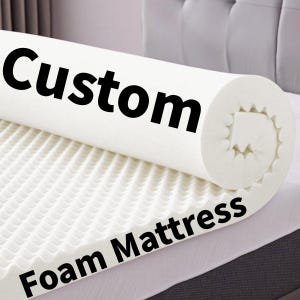 May include: A white foam mattress topper with the word "Custom" printed in black on the rolled-up portion. The words "Foam Mattress" are printed in black on the unfolded portion. The topper has a textured surface.