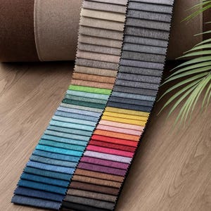May include: A collection of fabric swatches in various colors, including shades of blue, green, red, and brown. The samples are arranged in rows, showcasing different textures and hues. The image also includes a rolled-up fabric and a green plant.