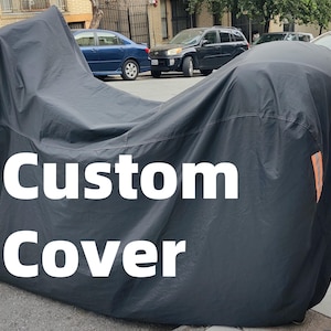 May include: A black custom cover draped over a motorcycle, with the words "Custom Cover" in white text. The cover is outdoors, parked on a city street.