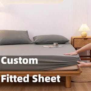Può includere: Un lenzuolo con angoli grigio su un letto con struttura in legno, con due cuscini abbinati. Il testo "Custom Fitted Sheet" è visualizzato sul lenzuolo. Un piccolo comodino con una lampada è visibile sullo sfondo.