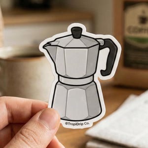 May include: A die-cut sticker featuring a silver moka pot with a black handle and knob. The design has a thick white border and the text TropiDrip Co. printed in small black letters at the bottom center of the sticker's base.