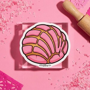 May include: A pink and gold concha sticker with a white border, set on a clear acrylic block. The sticker features a detailed illustration of the traditional Mexican sweet bread. The background is a solid pink color, with a wooden rolling pin in the upper right corner. The sticker has the text "©TropiDrip Co."