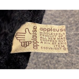 May include: Close-up of a tag from the brand "applause" with a hand logo. The tag is beige with brown text and is attached to a dark, fuzzy material. The text includes "Middlesex, N.J." and "Copyright".