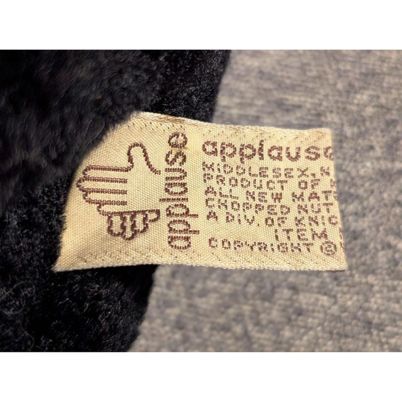 May include: Close-up of a tag from the brand "applause" with a hand logo. The tag is beige with brown text and is attached to a dark, fuzzy material. The text includes "Middlesex, N.J." and "Copyright".
