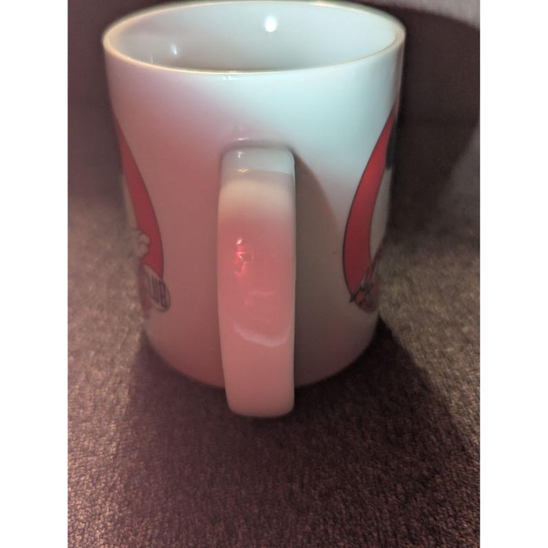 May include: White ceramic mug with a curved handle. The mug features a red and black design with text. The mug is cylindrical and appears to be clean.