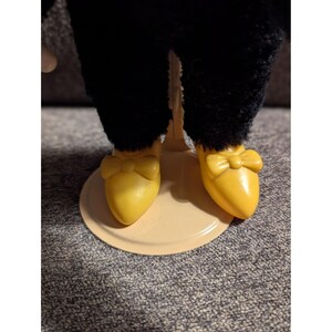 May include: A close-up of a doll's legs and feet. The doll wears black pants and yellow shoes with bow accents. The doll is standing on a light tan circular base.