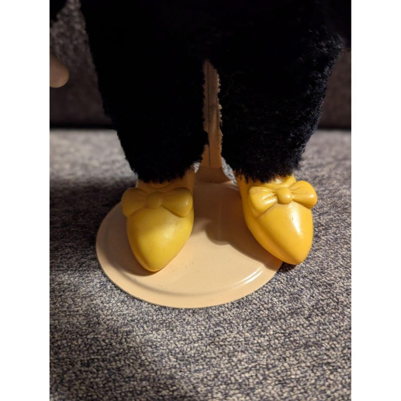 May include: A close-up of a doll's legs and feet. The doll wears black pants and yellow shoes with bow accents. The doll is standing on a light tan circular base.