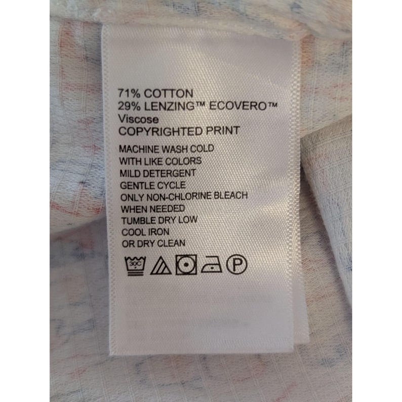 May include: A white fabric care label with black text reading 71% COTTON, 29% LENZING ECOVERO VISCOSE, and COPYRIGHTED PRINT. It includes care instructions for machine washing, non-chlorine bleach, tumble drying, and ironing, plus five laundry symbols.