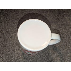 May include: White ceramic coffee mug with a curved handle. The bottom of the mug is visible, showing a white surface. The mug has a red and white design around the base.