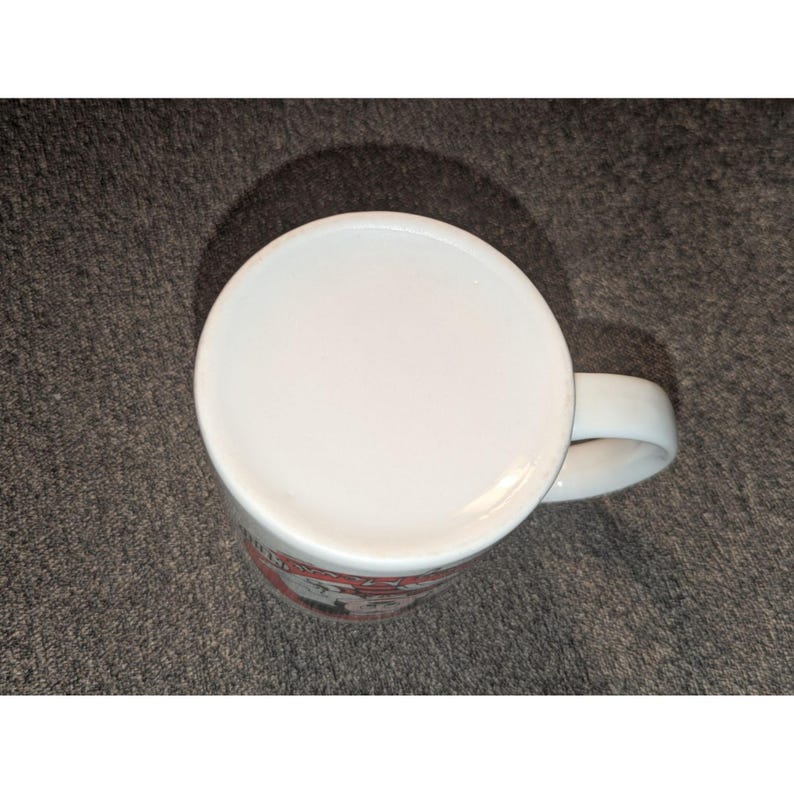 May include: White ceramic coffee mug with a curved handle. The bottom of the mug is visible, showing a white surface. The mug has a red and white design around the base.