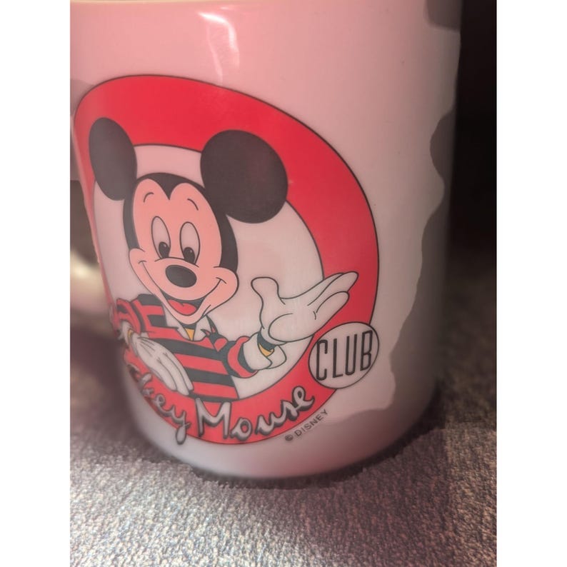 May include: White ceramic mug featuring Mickey Mouse from the Mickey Mouse Club. The design includes Mickey in a red, black, and white outfit, set against a red circle. The words "Mickey Mouse Club" and "Disney" are also visible.