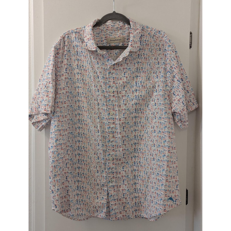May include: A short-sleeved button-down shirt in white featuring an all-over print of small, colorful cocktail glasses and tropical drink icons in shades of blue, orange, and tan. The shirt has a standard collar and a single chest pocket.