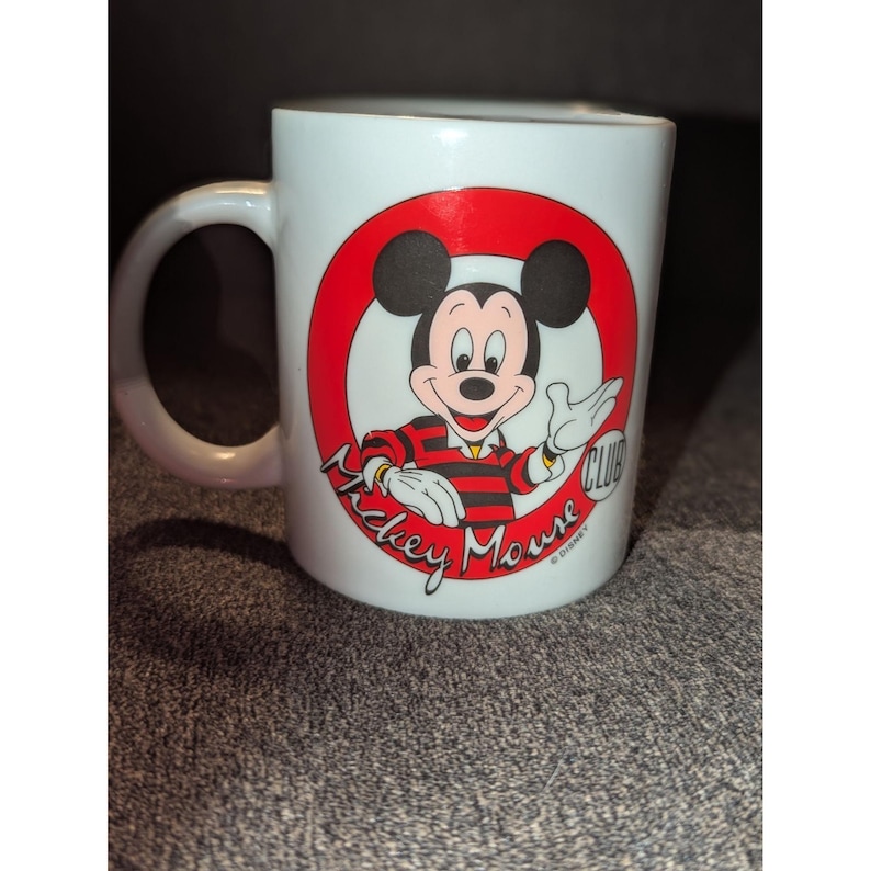 May include: White ceramic mug featuring Mickey Mouse in a red circle with the words "Mickey Mouse Club". Mickey is wearing a red and black striped shirt. The mug has a white handle and is a classic collectible.