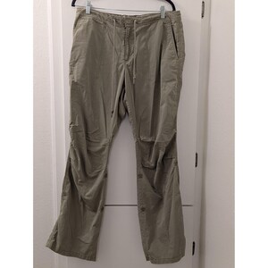 May include: Olive green pants with a drawstring waist and button details. The pants have a relaxed fit and feature pockets on the sides. The legs have button closures at the bottom. The pants are hanging on a hanger.