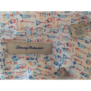 May include: A white button-down shirt featuring a repeating pattern of colorful cocktail glasses and drink accessories in blue, orange, and red. A beige Tommy Bahama brand label is stitched on the interior, with a size XL tag reading Made in Vietnam.