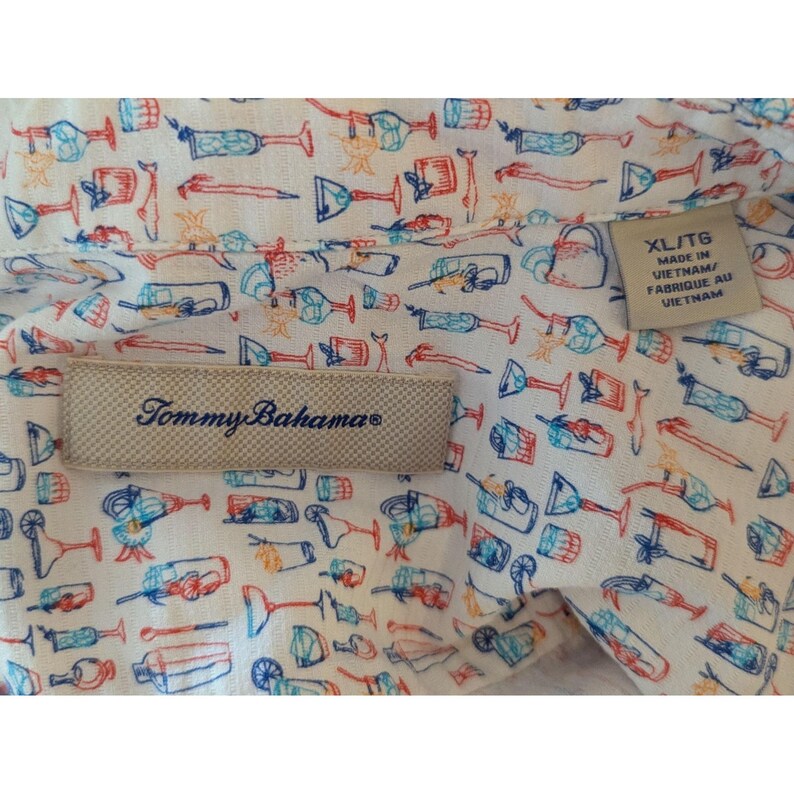 May include: A white button-down shirt featuring a repeating pattern of colorful cocktail glasses and drink accessories in blue, orange, and red. A beige Tommy Bahama brand label is stitched on the interior, with a size XL tag reading Made in Vietnam.