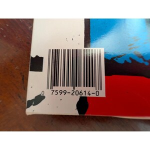 Puede incluir: Primer plano de una esquina de producto con un c&oacute;digo de barras y el n&uacute;mero "0 7599-20614-0" impreso debajo. El producto tiene un fondo blanco con detalles en negro, rojo y azul.