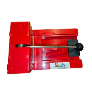 May include: A bright red Sizzix die-cutting machine featuring a rectangular base, a metal handle with a black grip, and a white label with the Sizzix logo. The device has a sturdy plastic construction with a mechanical lever mechanism for operation.