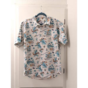 May include: White button-down shirt with a colorful desert-themed print. The short-sleeved shirt features illustrations of cacti, hot air balloons, and musical instruments. The shirt has the words "Coachella", "Indio", and "Mojave" printed on it.