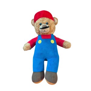 Può includere: Un orsacchiotto di peluche con pelliccia marrone chiaro, che indossa un berretto rosso, una maglietta rossa a maniche lunghe e una salopette blu con due bottoni gialli. L'orso ha baffi neri ricamati, occhi blu e piedi marrone scuro.