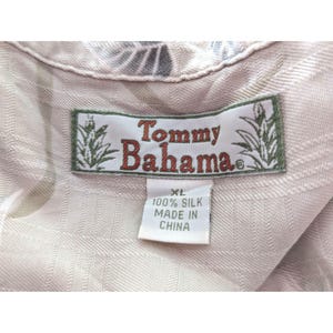 Może przedstawiać: Jasnobeżowa koszula jedwabna z metką marki Tommy Bahama z czerwonym napisem i grafiką ananasa. Biała metka pod marką wskazuje rozmiar XL, skład materiałowy 100% jedwab oraz kraj produkcji Chiny.