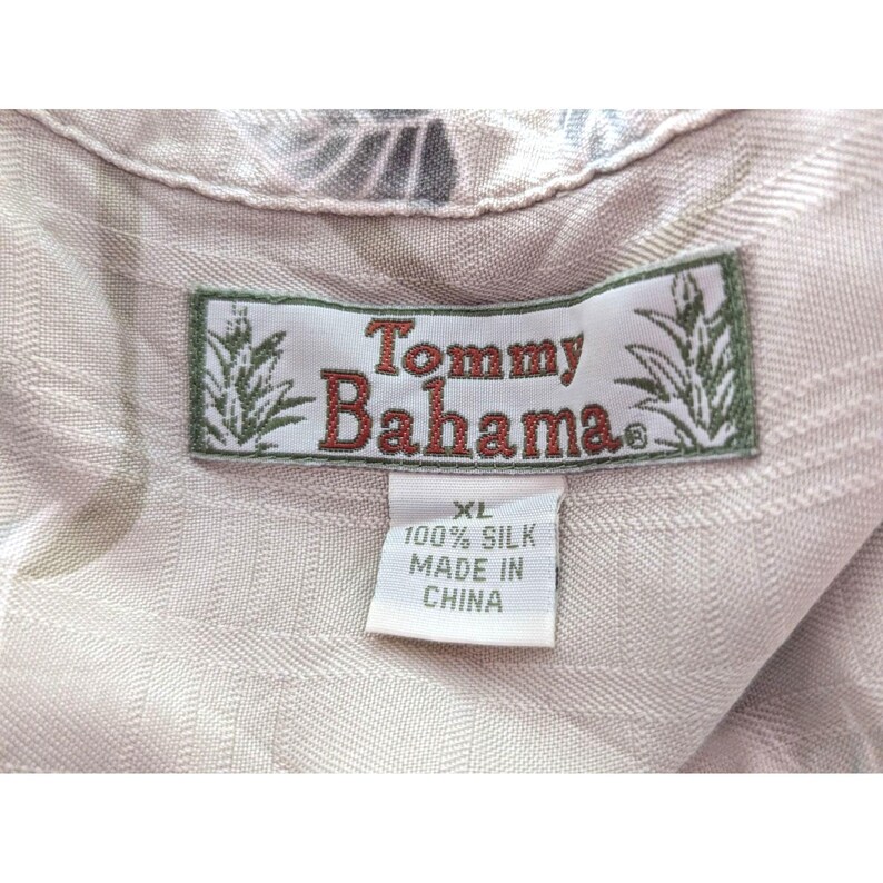 Może przedstawiać: Jasnobeżowa koszula jedwabna z metką marki Tommy Bahama z czerwonym napisem i grafiką ananasa. Biała metka pod marką wskazuje rozmiar XL, skład materiałowy 100% jedwab oraz kraj produkcji Chiny.