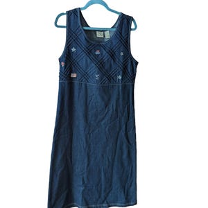 May include: A sleeveless denim dress with a scoop neckline. The bodice features a diamond-stitched pattern with embroidered floral and star accents. An American flag patch is visible on the bodice. The dress hangs on a blue hanger.