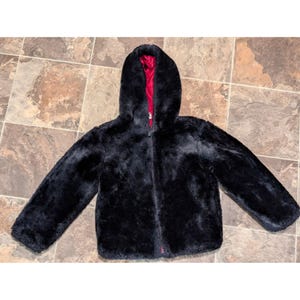 May include: A black faux fur jacket with a hood and a red interior lining. The jacket has a zipper closure and long sleeves. The jacket is laying flat on a tiled surface.