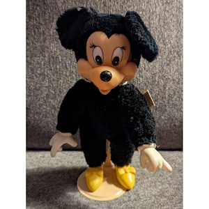 May include: A vintage Minnie Mouse plush doll with a black fuzzy body, large ears, and a tan face. The doll has white gloves, yellow shoes, and is standing on a beige stand. The doll has a tag attached.