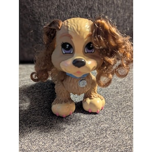 May include: A tan and brown puppy figurine with curly brown hair and purple eyes. The puppy wears a blue collar with a silver tag. The paws are tan with pink pads. The toy is on a gray surface.