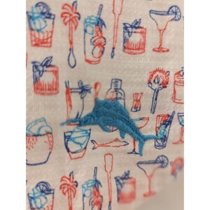 May include: A white fabric featuring a repeating pattern of red and blue line-drawn cocktail glasses, palm trees, and bar tools, with a prominent, textured light blue embroidered marlin fish centered over the printed cocktail illustrations.