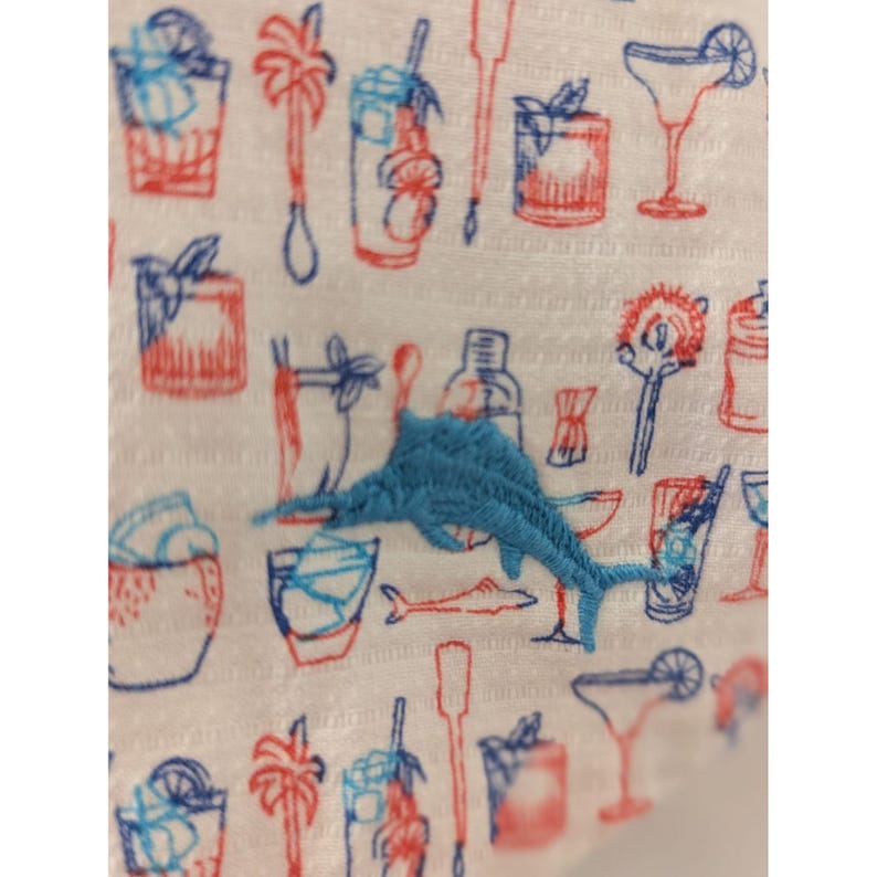 May include: A white fabric featuring a repeating pattern of red and blue line-drawn cocktail glasses, palm trees, and bar tools, with a prominent, textured light blue embroidered marlin fish centered over the printed cocktail illustrations.