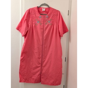 May include: A coral-colored, short-sleeved robe with a textured pattern and a full-length zipper. The robe features embroidered floral details in shades of blue and green near the neckline. It is hanging on a gray hanger.